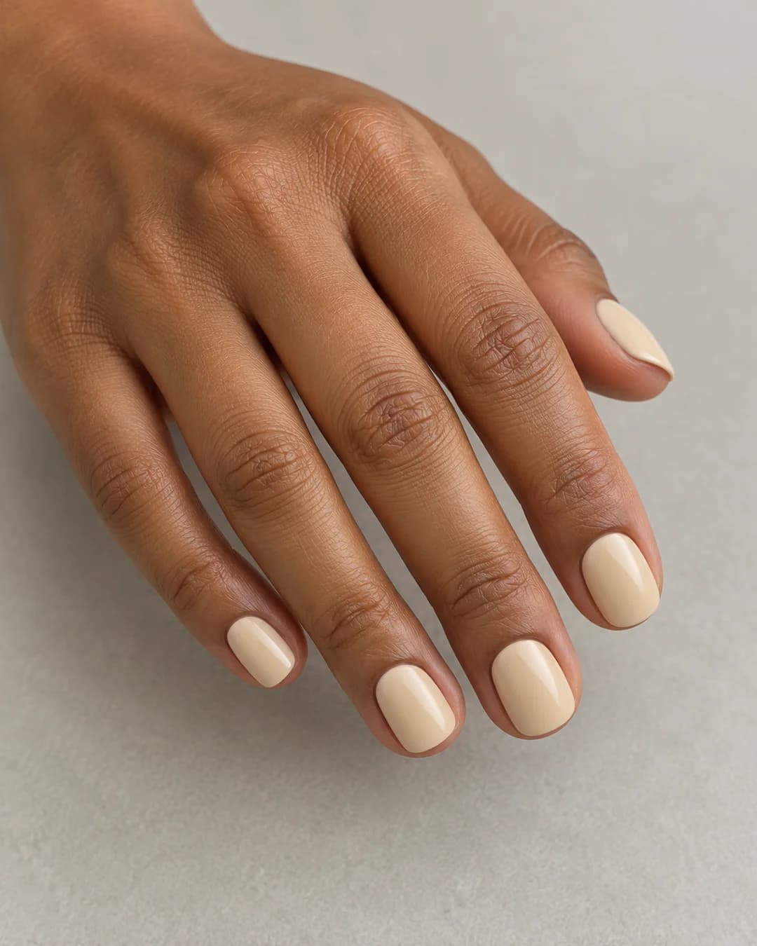 Uñas cortas cuadradas de bordes redondeados de color beige cálido cremoso uniforme y acabado satinado. Vista oblicua de la mano.