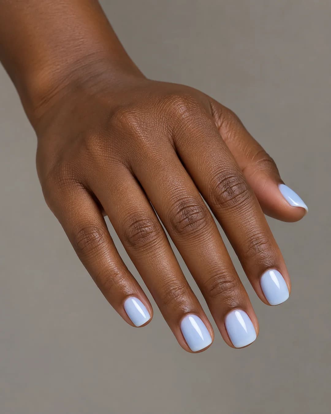Uñas de forma cuadrada y longitud corta, en azul pastel lechoso de acabado brillante, con esquinas suaves. Vista oblicua de la mano.