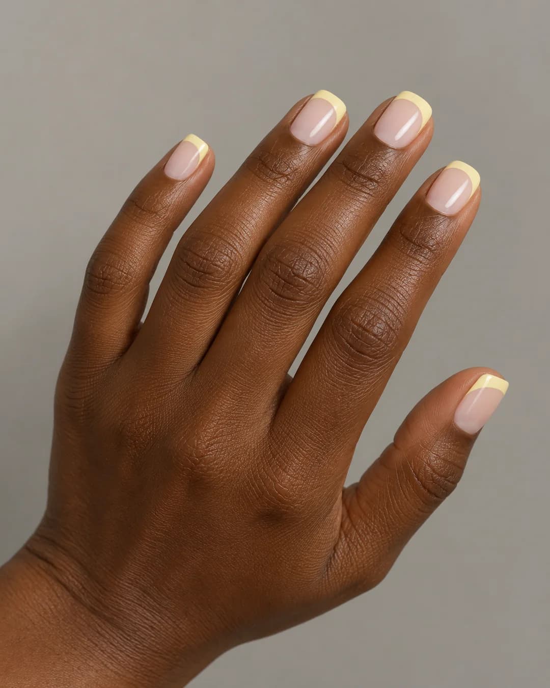 Uñas de forma cuadrada y longitud corta, en base lechosa translúcida y puntas francesas amarillo mantequilla, con esquinas suaves. Vista oblicua de la mano.