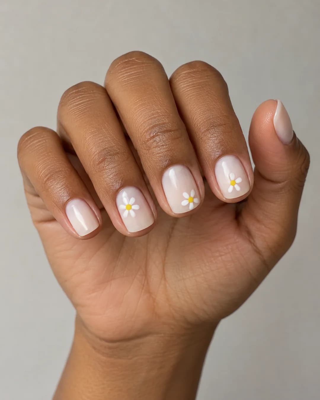 Uñas cortas cuadradas de bordes redondeados con base blanca lechosa y pequeñas margaritas blancas de centro amarillo. Vista oblicua de la mano.
