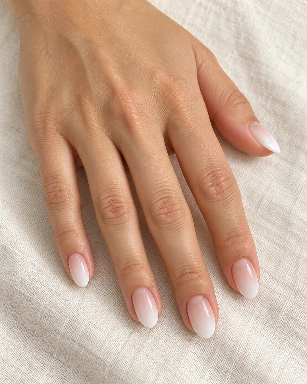 Uñas cortas almendradas con degradado suave de nude cálido a blanco lechoso y acabado satinado. Detalle de las uñas.