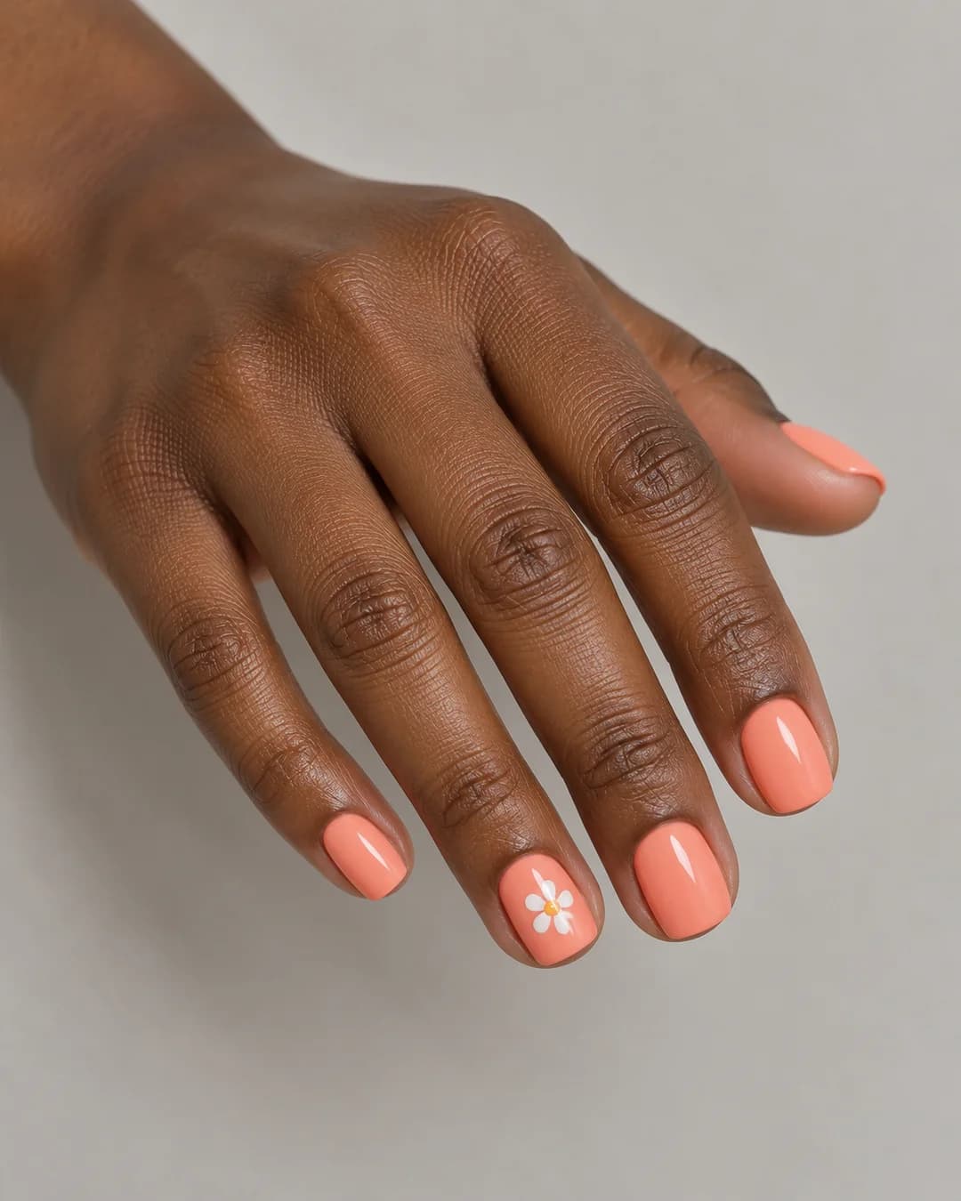 Uñas cortas cuadradas de bordes redondeados de color coral melocotón satinado y una margarita blanca en la uña del anular. Vista oblicua de la mano.