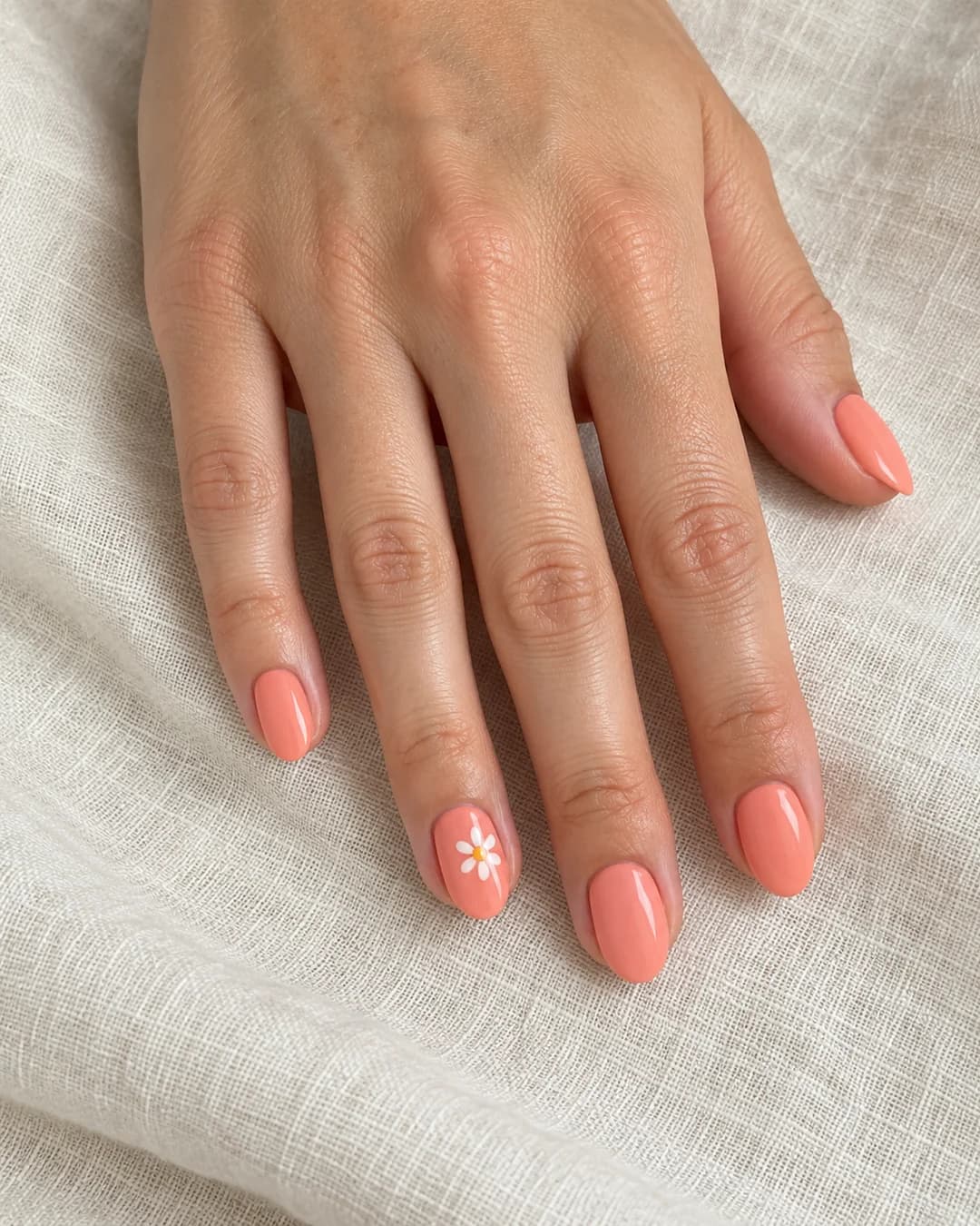 Uñas cortas ovaladas de color coral melocotón satinado y una margarita blanca en la uña del anular. Detalle de las uñas.