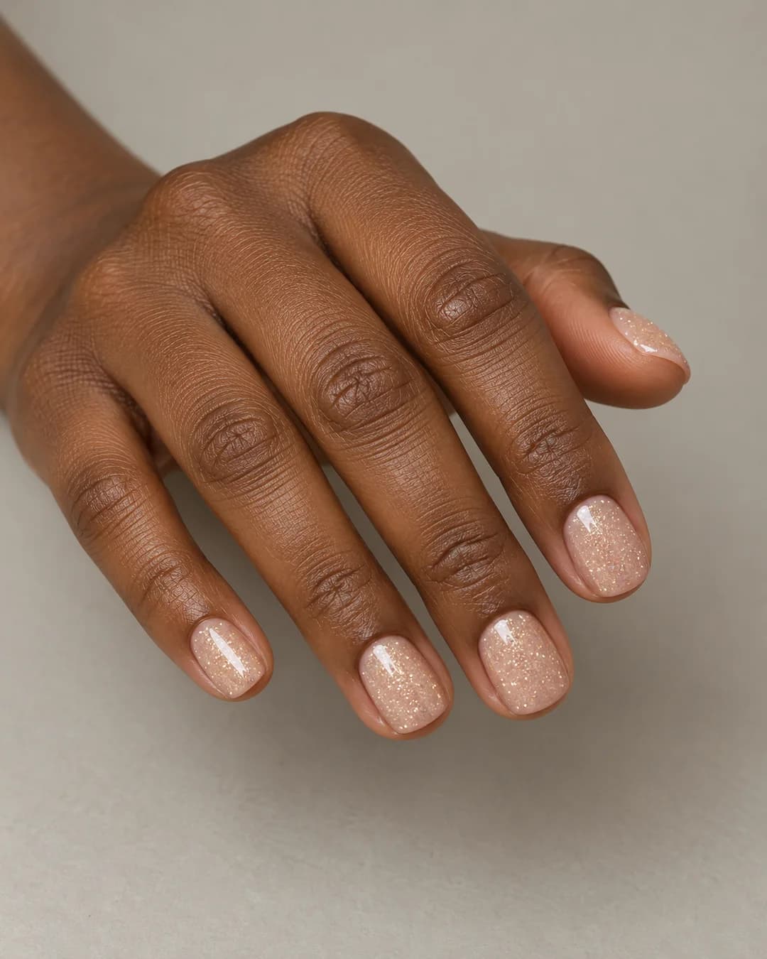 Uñas cortas cuadradas de bordes redondeados con base nude y purpurina fina de color dorado champán. Vista oblicua de la mano.