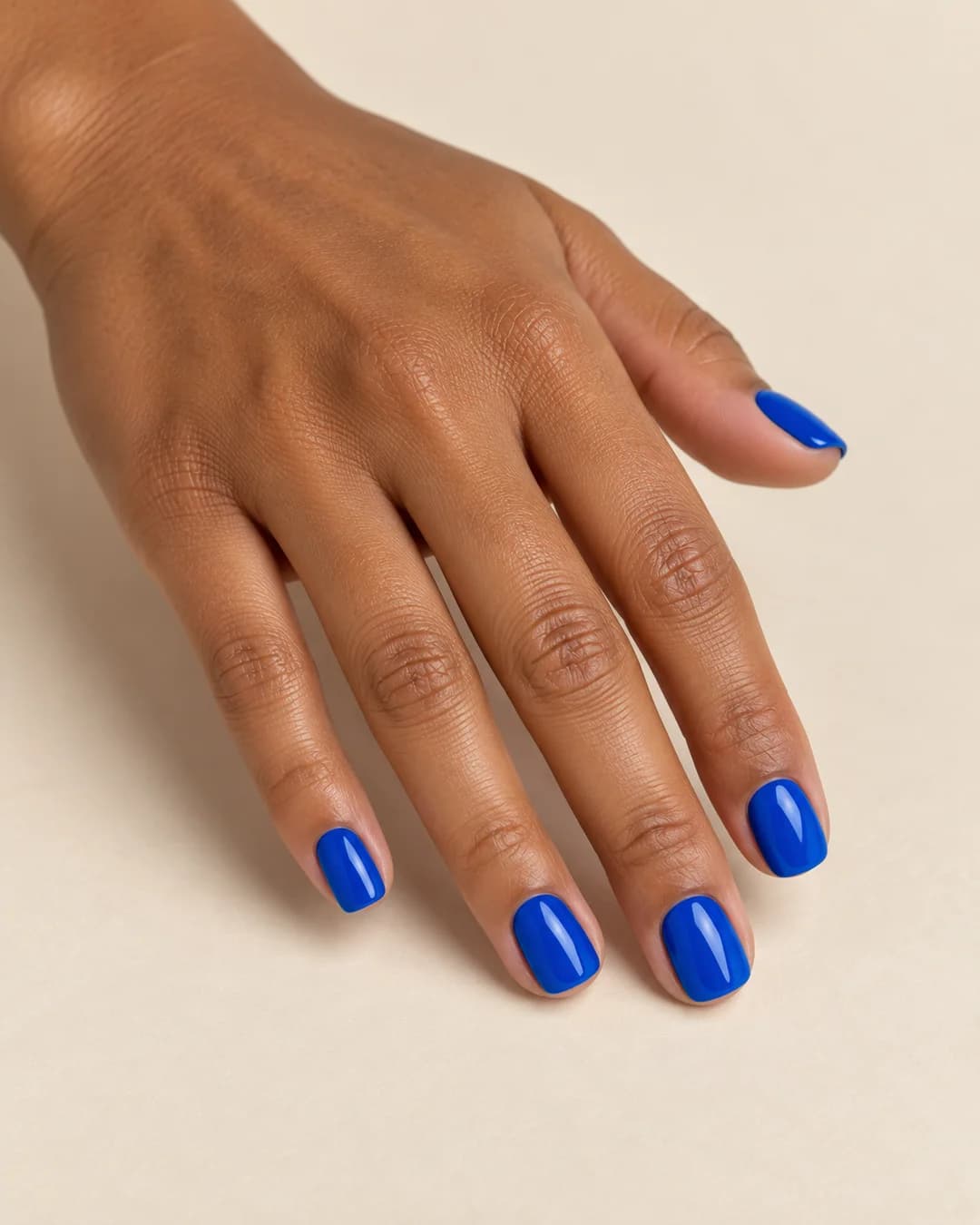 Mano de piel morena con uñas cortas cuadradas de bordes suaves en azul cobalto brillante. Vista oblicua de la mano.