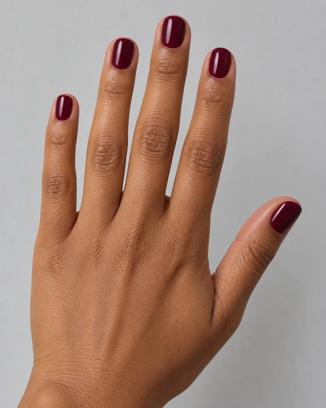 Mano de piel morena con uñas cortas redondeadas en burdeos brillante. Detalle de las uñas.