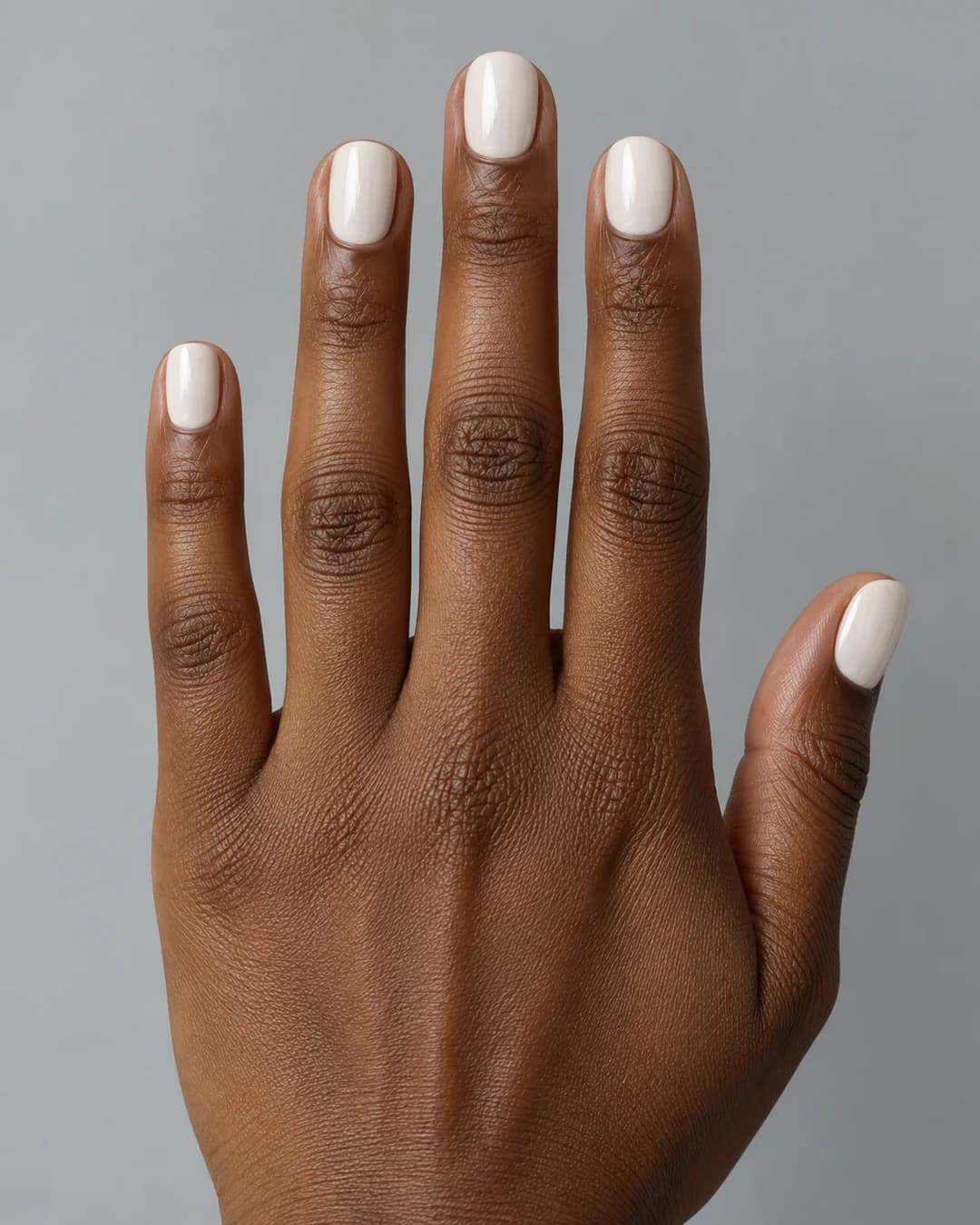 Mano de piel oscura con uñas cortas redondeadas en blanco roto brillante. Detalle de las uñas.
