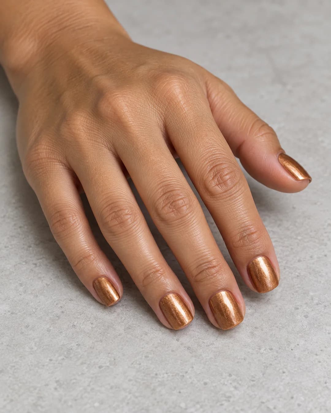 Mano de piel tostada con uñas cortas cuadradas de bordes suaves en dorado cobrizo metálico. Vista oblicua de la mano.