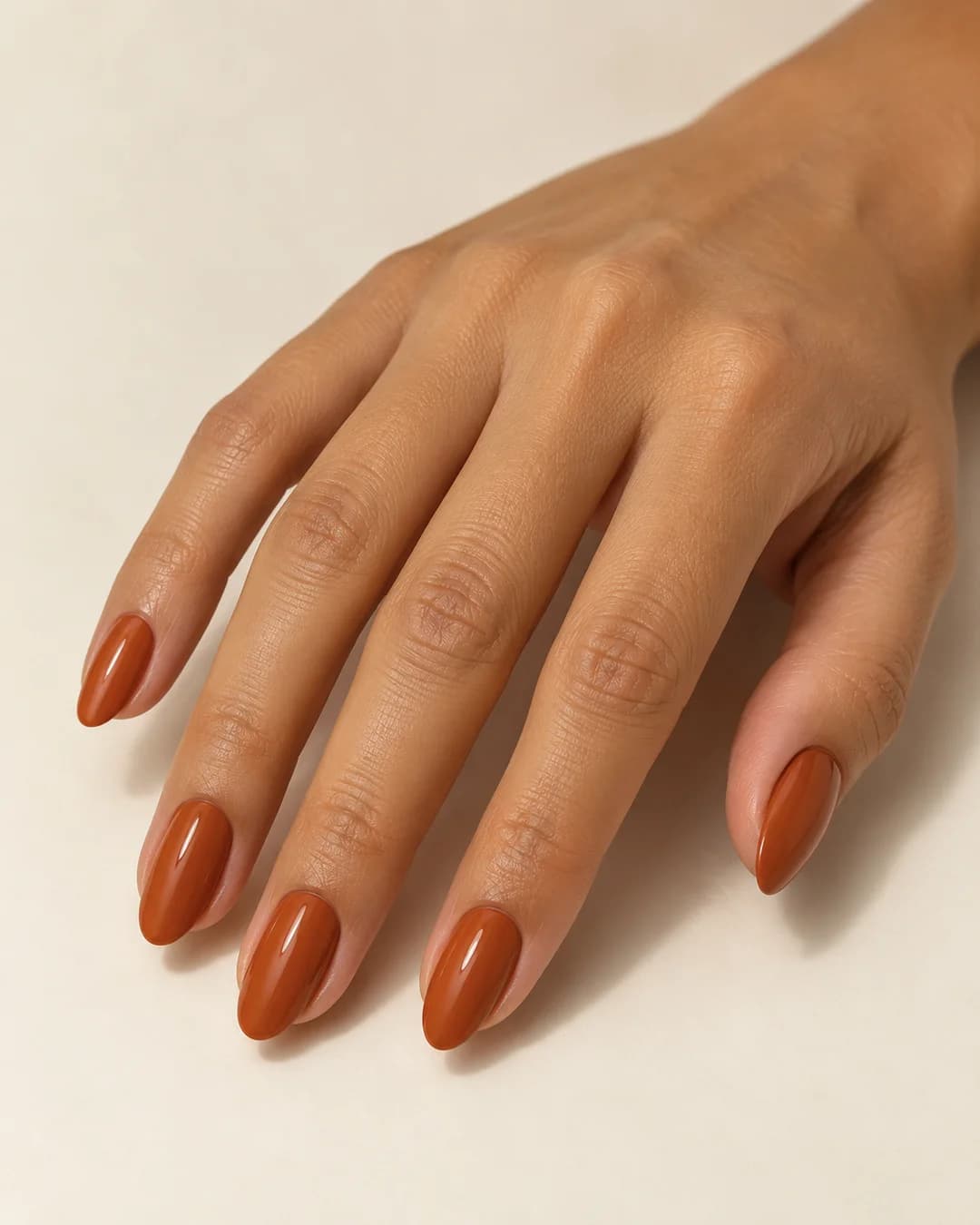 Mano de piel tostada con uñas almendradas de largo medio en terracota brillante. Vista oblicua de la mano.