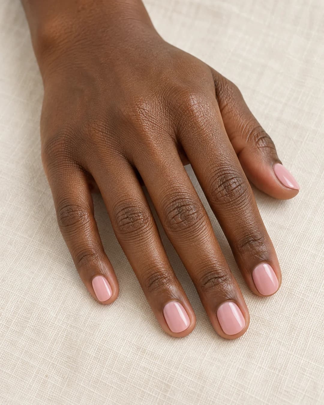 Mano adulta de piel marrón profunda con uñas cortas redondas en rosa empolvado brillante.