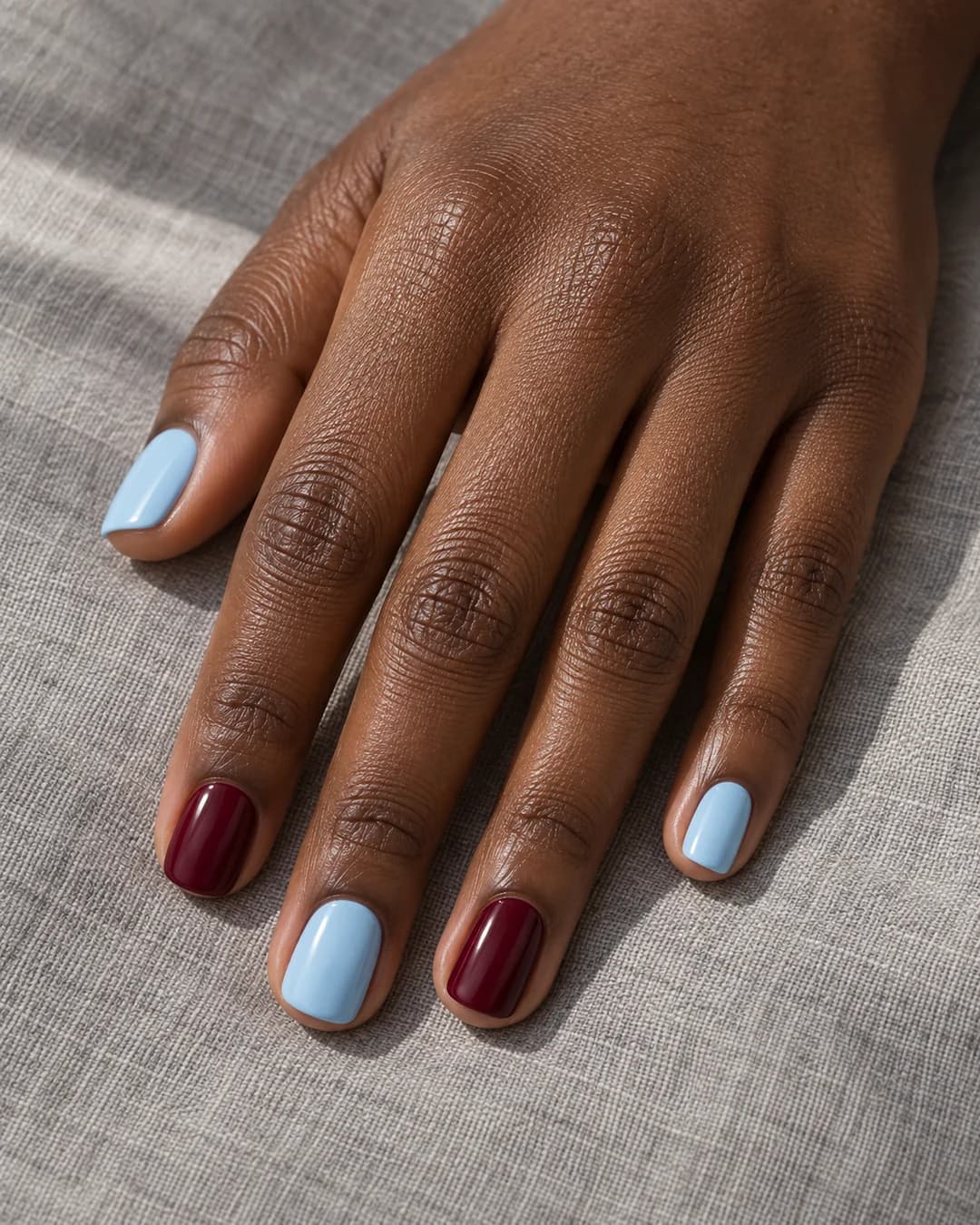 Mano de piel marrón oscura con uñas squoval cortas alternando esmalte liso azul bebé y burdeos.