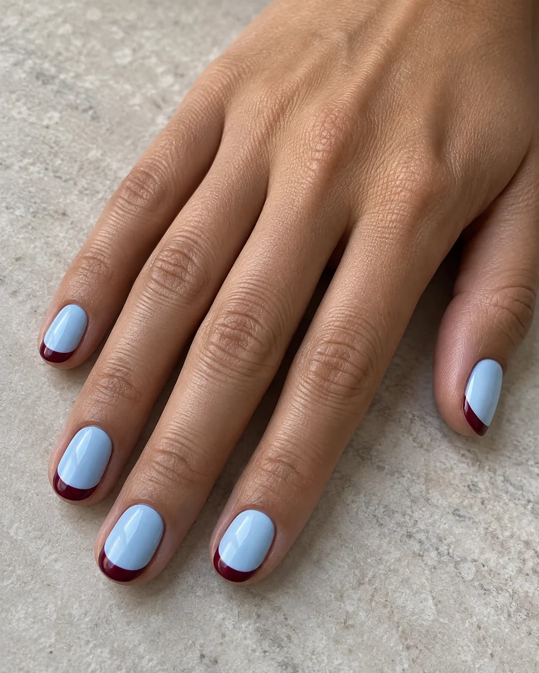Mano de piel oliva clara con uñas ovaladas cortas en azul bebé con puntas francesas curvas burdeos.