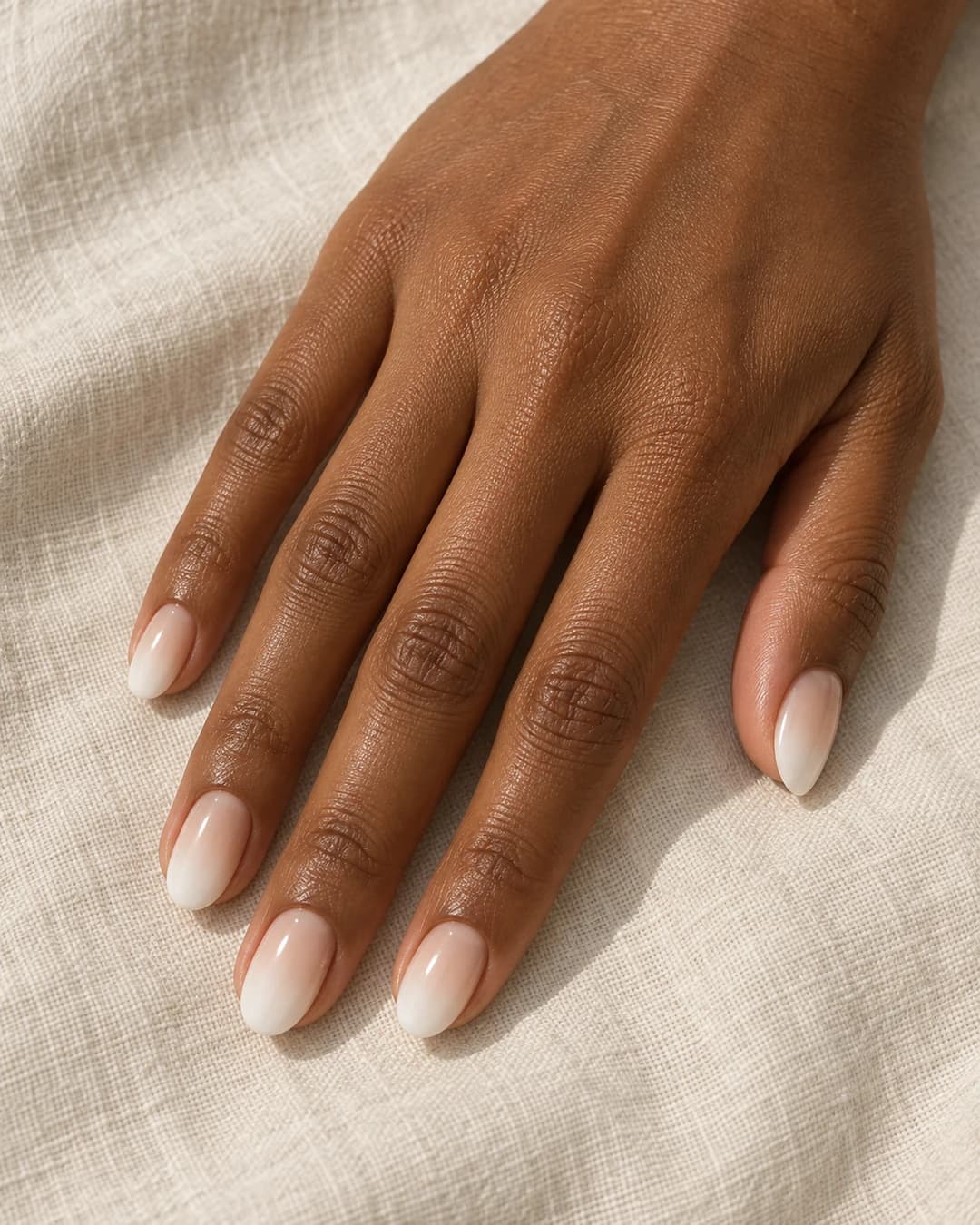 Uñas almendradas cortas con base crema translúcida y puntas blancas difuminadas sobre piel morena.