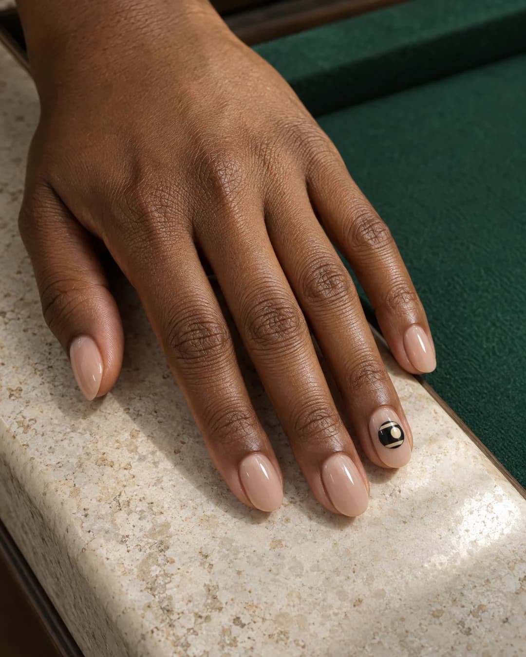 Mano adulta de piel marrón con uñas almendradas cortas sobre base nude translúcida y un pequeño motivo plano de bola negra de billar en la uña anular.