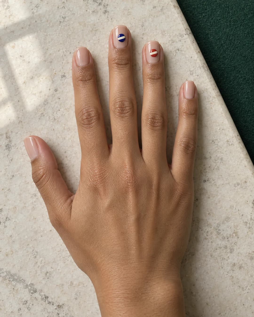 Mano adulta de piel oliva con cinco uñas squoval cortas en base nude translúcida y pequeños motivos planos de bolas de billar rayadas en azul cobalto y rojo.