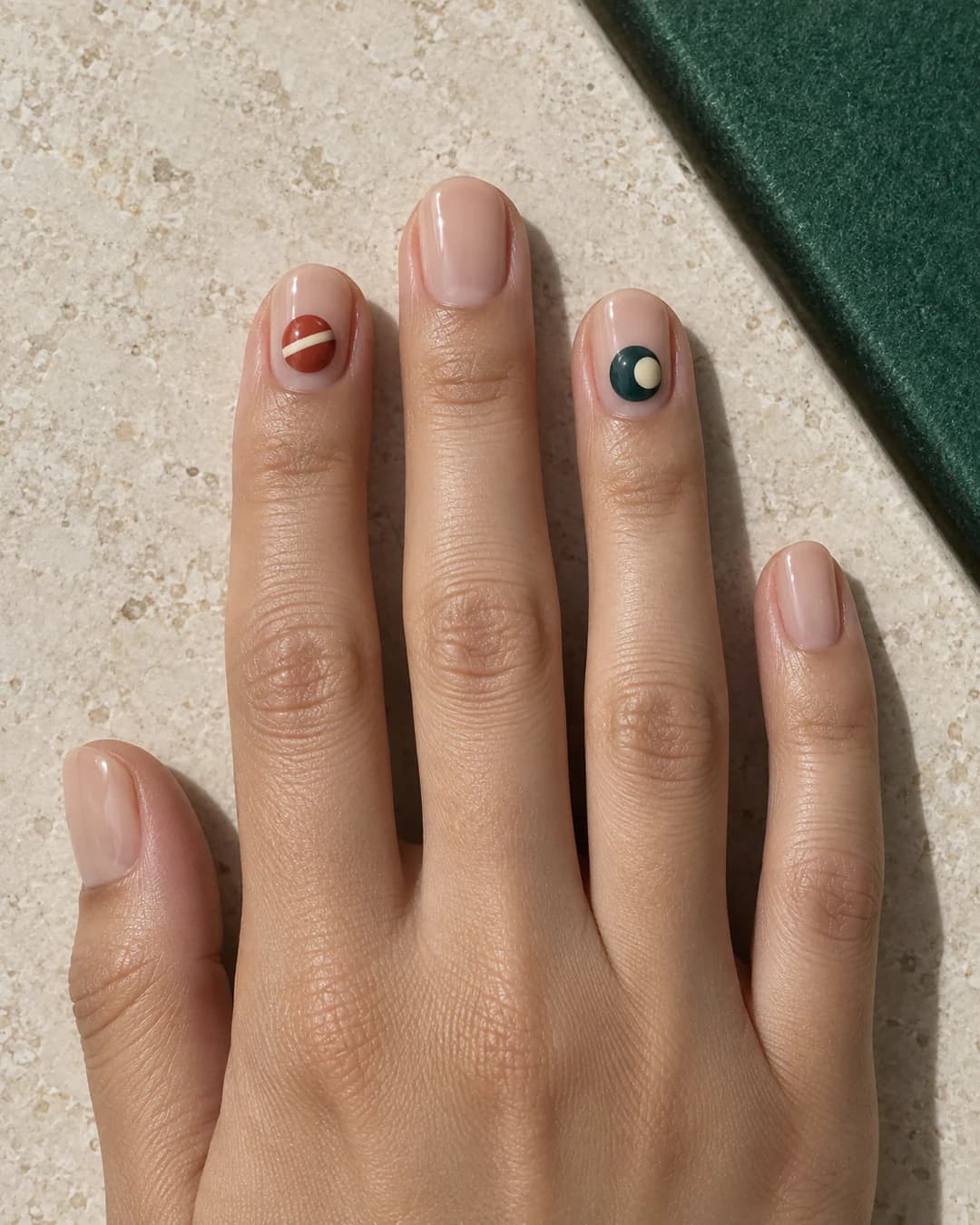 Mano adulta de piel marrón profunda con uñas ovaladas medias en base nude y pequeños motivos de bolas de billar terracota y azul petróleo.