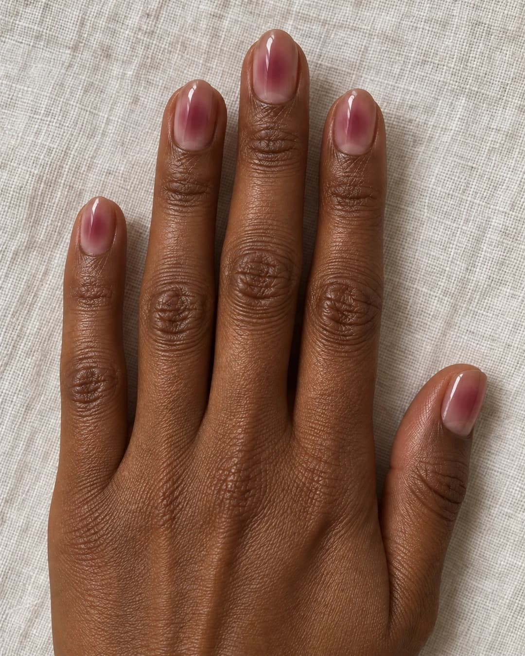 Mano adulta de piel marrón profunda con cinco uñas cortas almendradas en nude cacao translúcido y velo frambuesa malva difuminado.