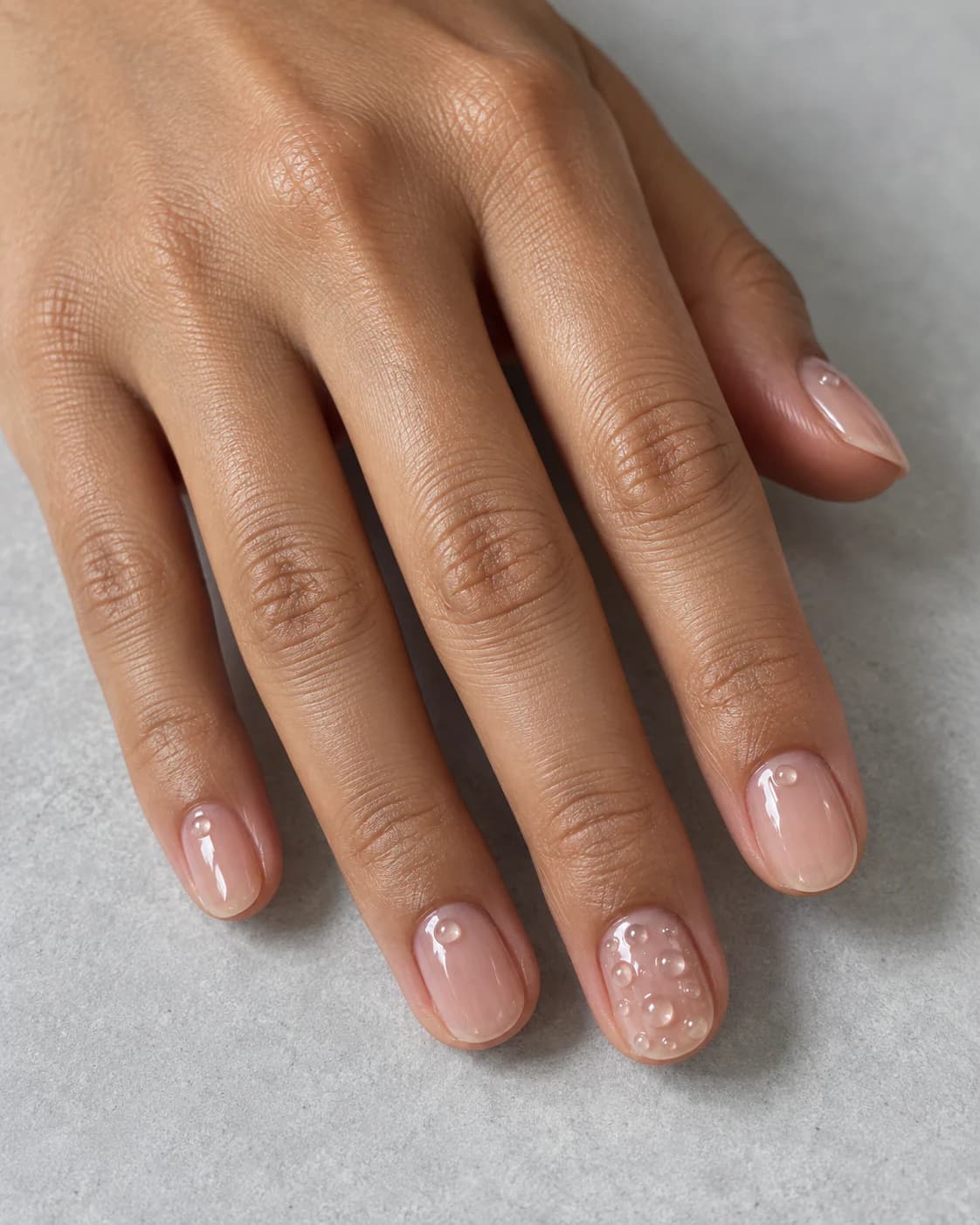 Mano adulta de piel morena clara con cinco uñas ovaladas cortas en jelly rosa translúcido y pequeñas burbujas de gel en el anular.