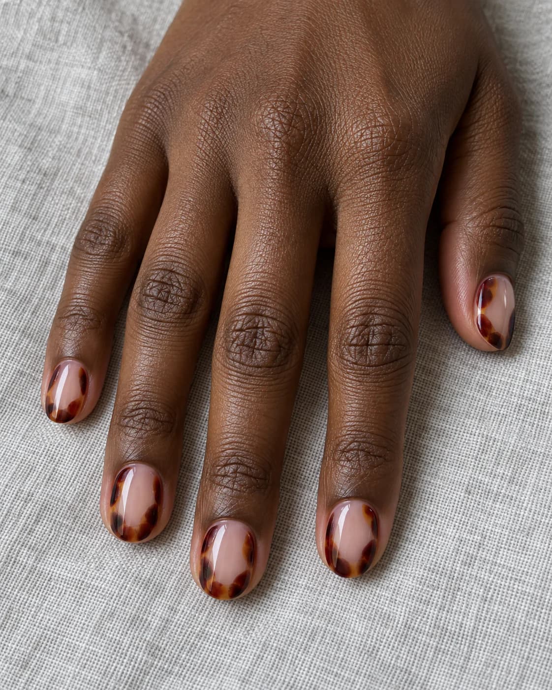 Mano adulta de piel marrón profunda con cinco uñas ovaladas cortas, paneles carey burdeos y base nude visible.