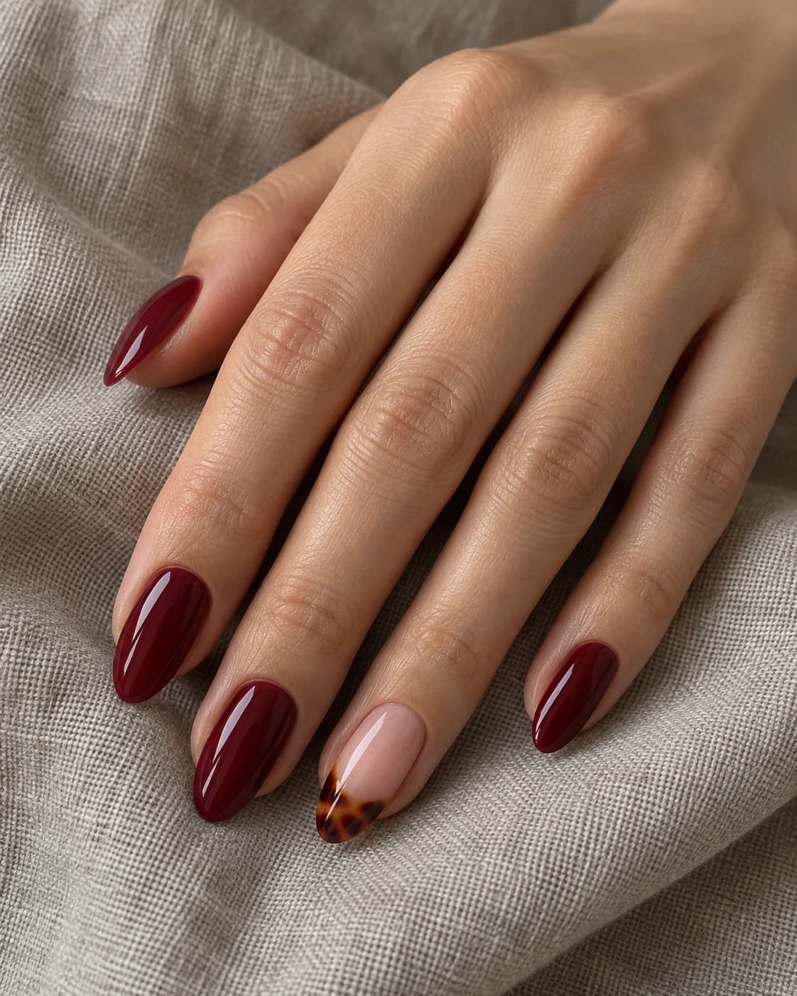 Mano adulta de piel clara con cuatro uñas almendradas en merlot y una punta carey translúcida en el anular.