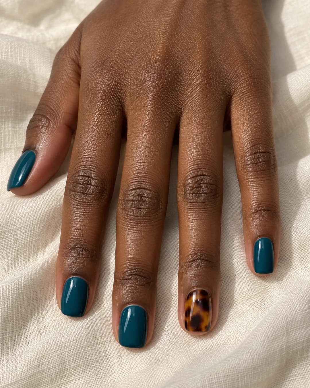 Mano de piel marrón profunda con uñas cortas cuadradas en turquesa petróleo y un anular carey ámbar y chocolate.