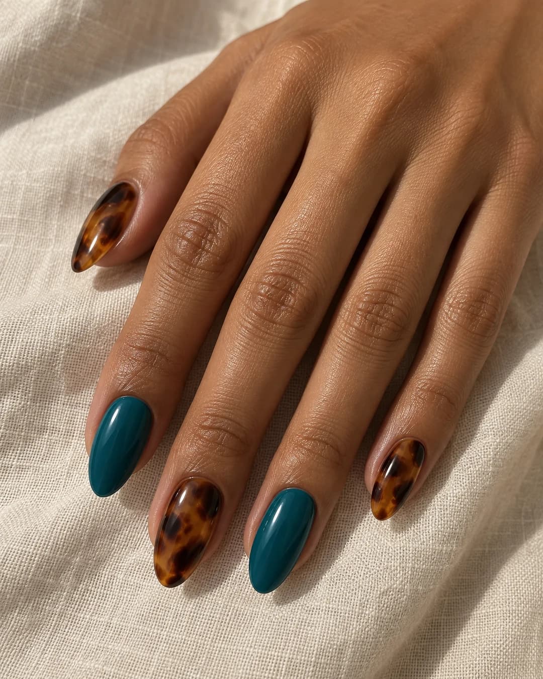 Mano de piel oliva media con uñas almendradas alternas en turquesa profundo y carey ámbar con chocolate.