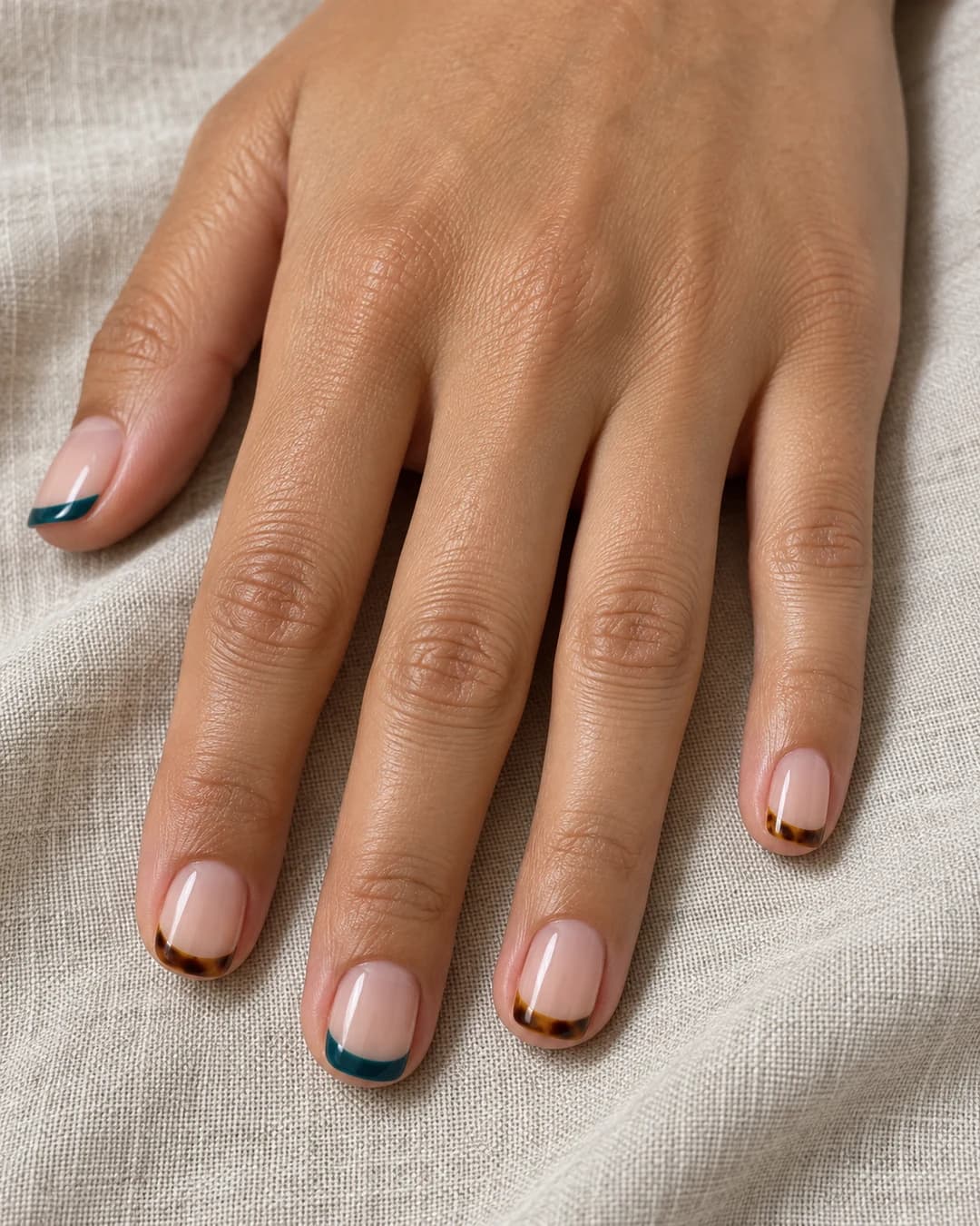 Mano de piel clara media con uñas squoval cortas de base nude y puntas finas alternas en turquesa y carey.