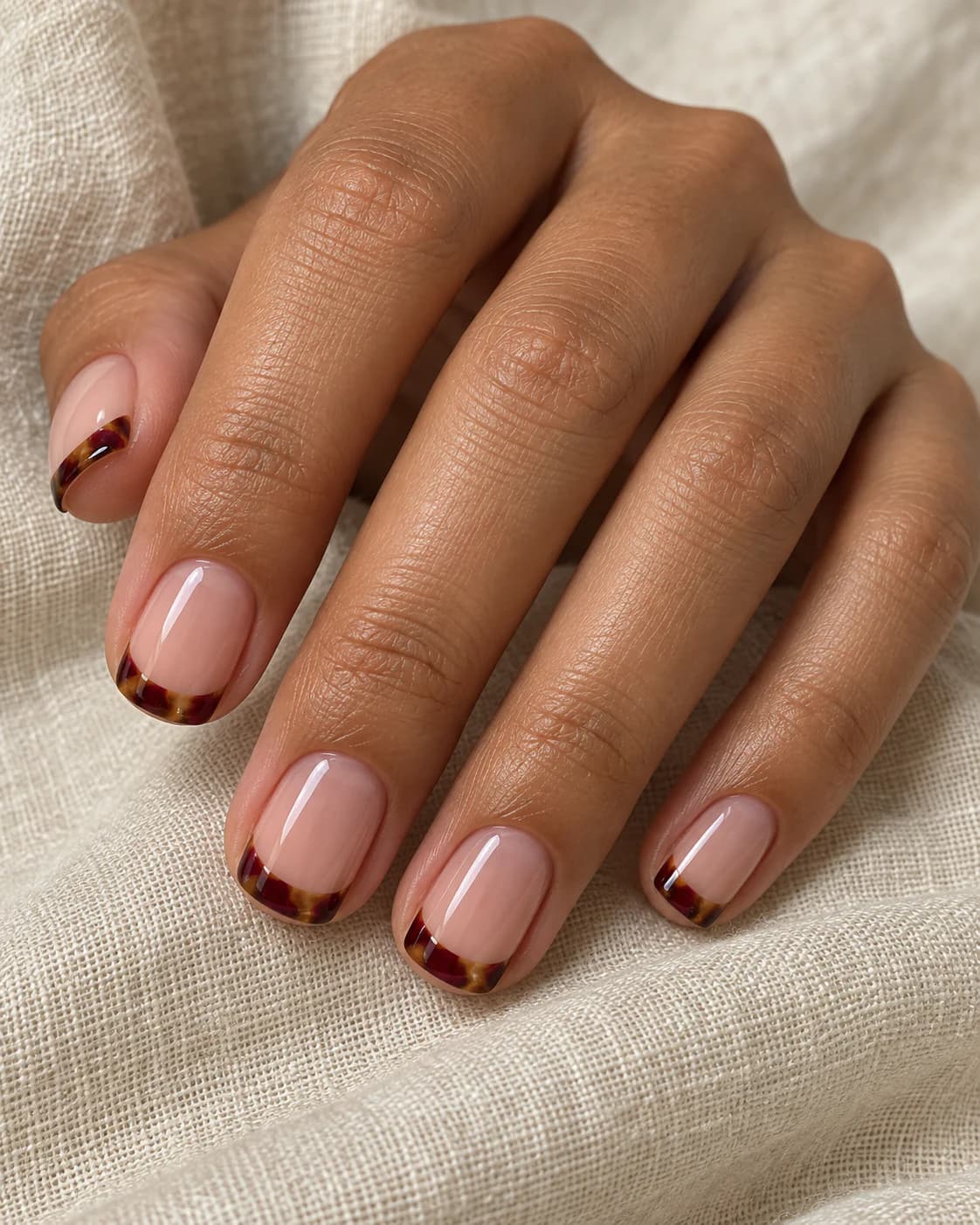 Mano adulta de piel oliva con cinco uñas squoval cortas y puntas carey translúcidas en rojo vino, ámbar y chocolate.