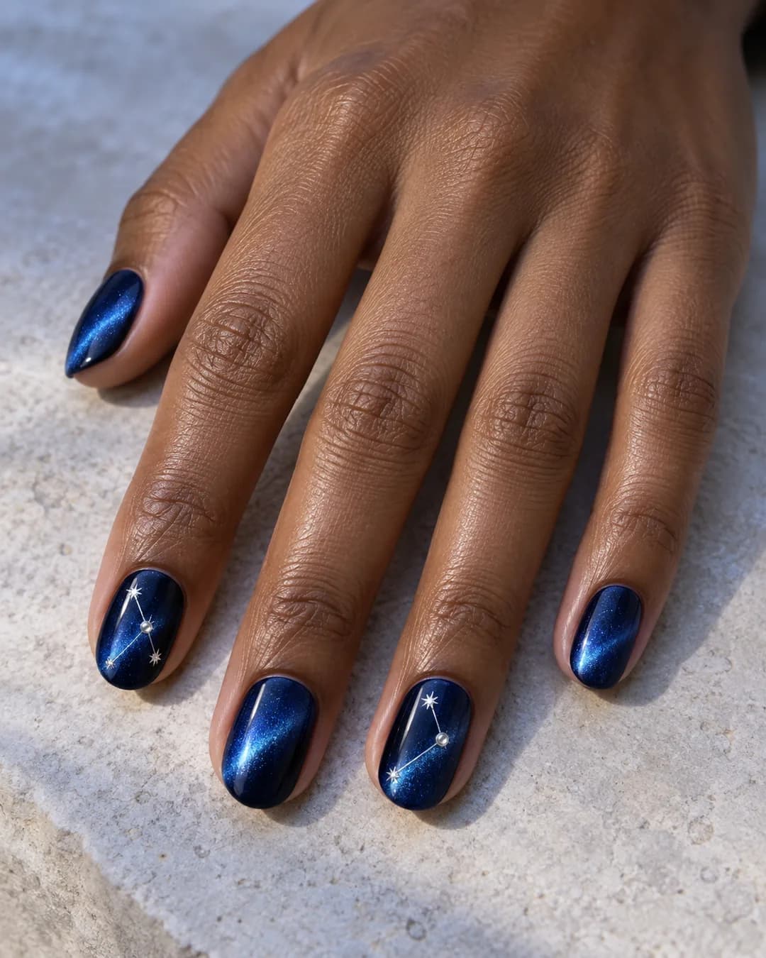 Mano adulta de piel marrón media con uñas ovaladas cortas cat-eye zafiro y finas constelaciones plateadas en dos acentos.