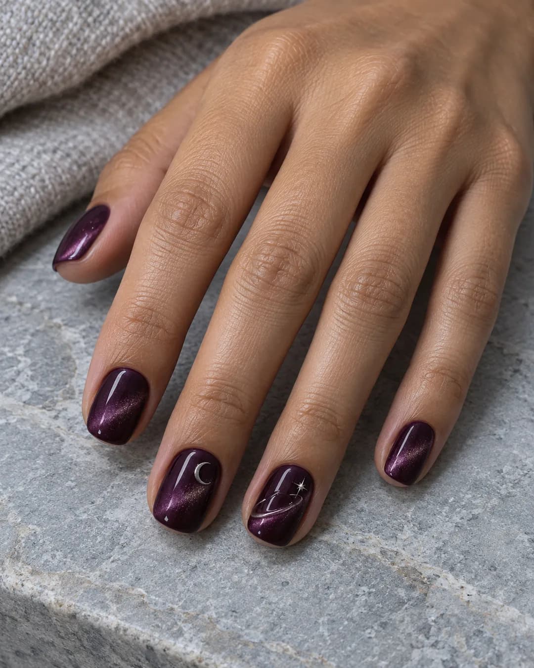 Mano adulta de piel oliva media con uñas squoval cortas en cat-eye ciruela y una luna plateada con órbita de gel transparente elevada en dos acentos.