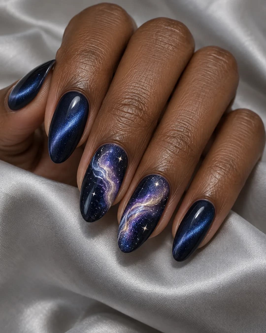 Mano adulta de piel marrón profunda con uñas almendradas medias en cat-eye índigo y dos acentos con Vía Láctea violeta, estrellas y gel transparente con relieve.