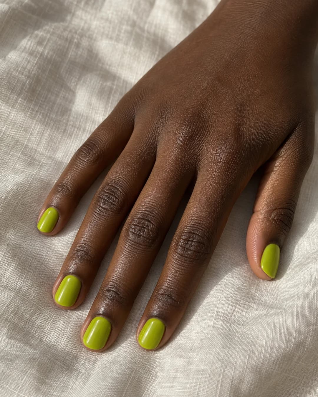 Mano de piel marrón profunda con uñas ovaladas cortas en chartreuse amarillo-verde brillante y acabado glossy.