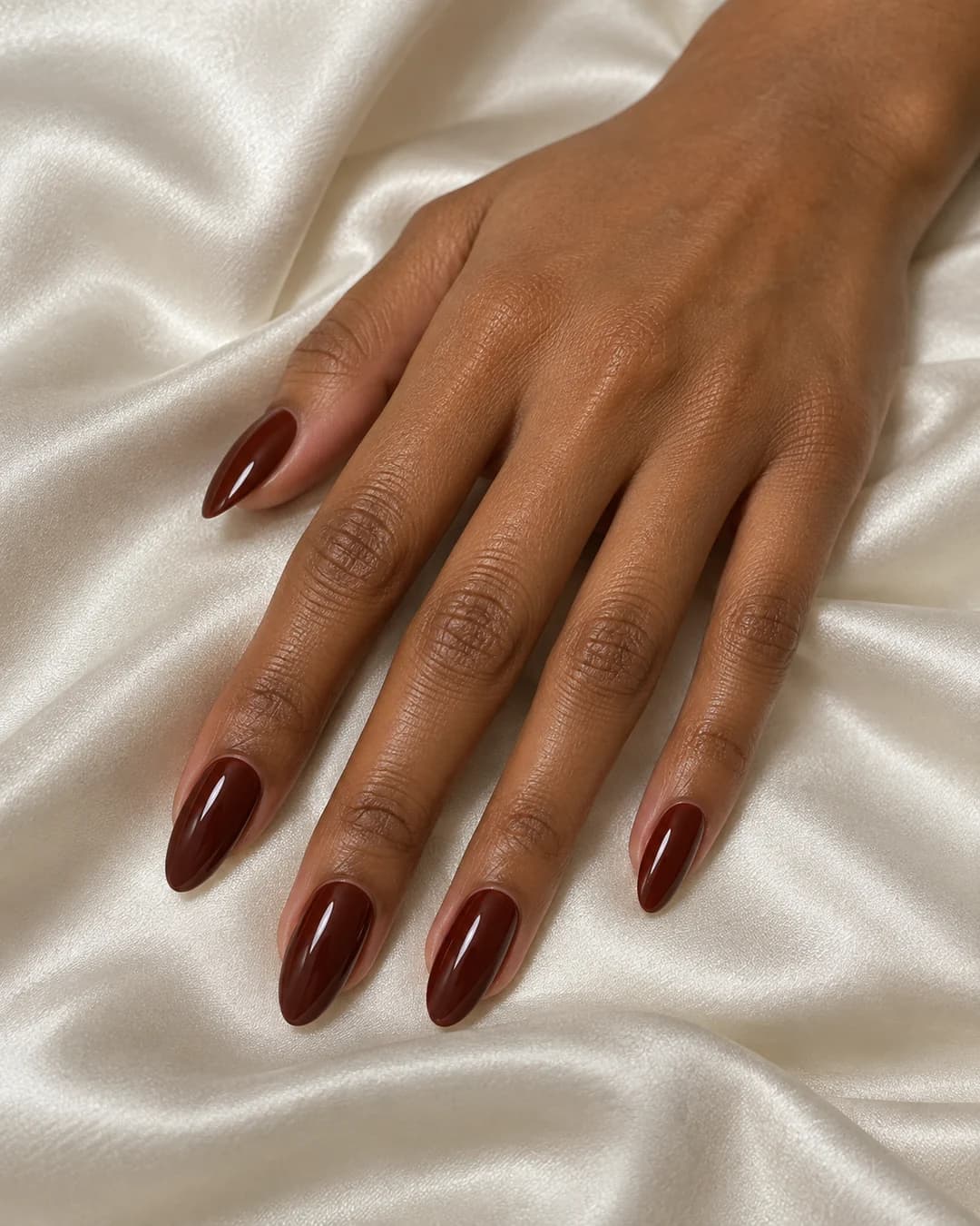 Mano adulta de piel marrón media con uñas almendradas medias en marrón rojizo chilli chocolate brillante.