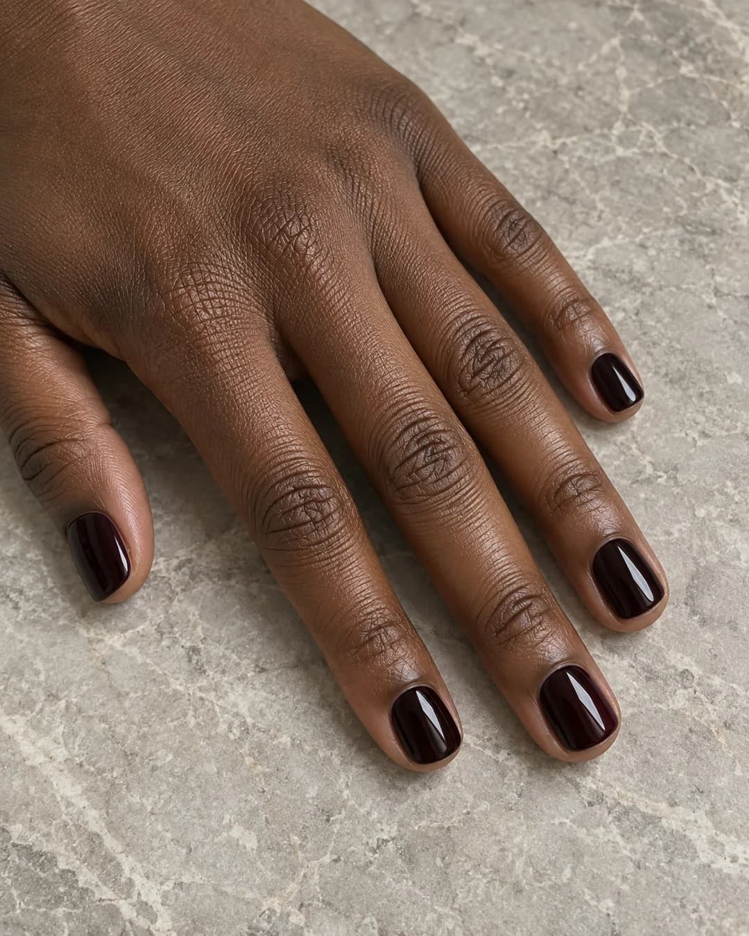 Mano adulta de piel marrón profunda con uñas cortas squoval en burdeos casi negro, estilo cereza oscura.