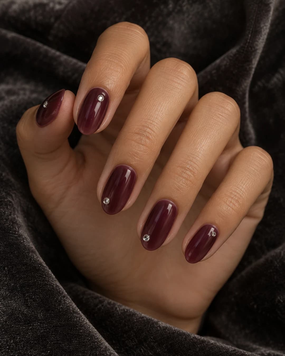 Mano adulta de piel oliva media con cinco uñas almendradas cortas color vino jelly y diminutos cristales transparentes en varias uñas.