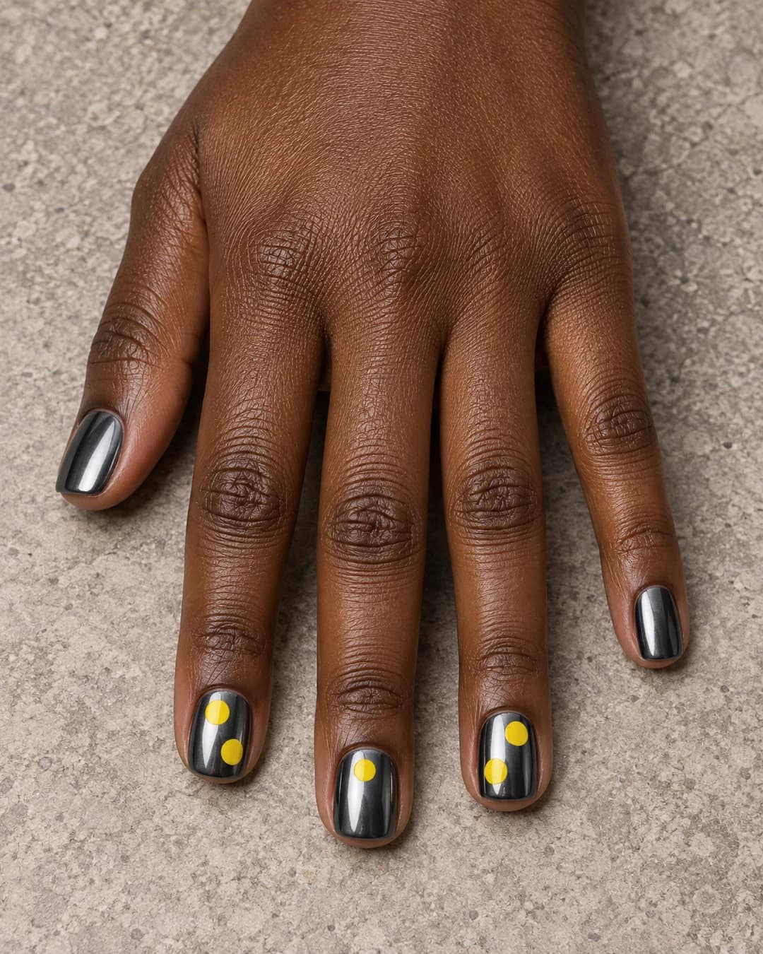 Mano adulta de piel marrón profunda con uñas squoval cortas en cromo gunmetal, puntos amarillos grandes en tres uñas y dos lisas.