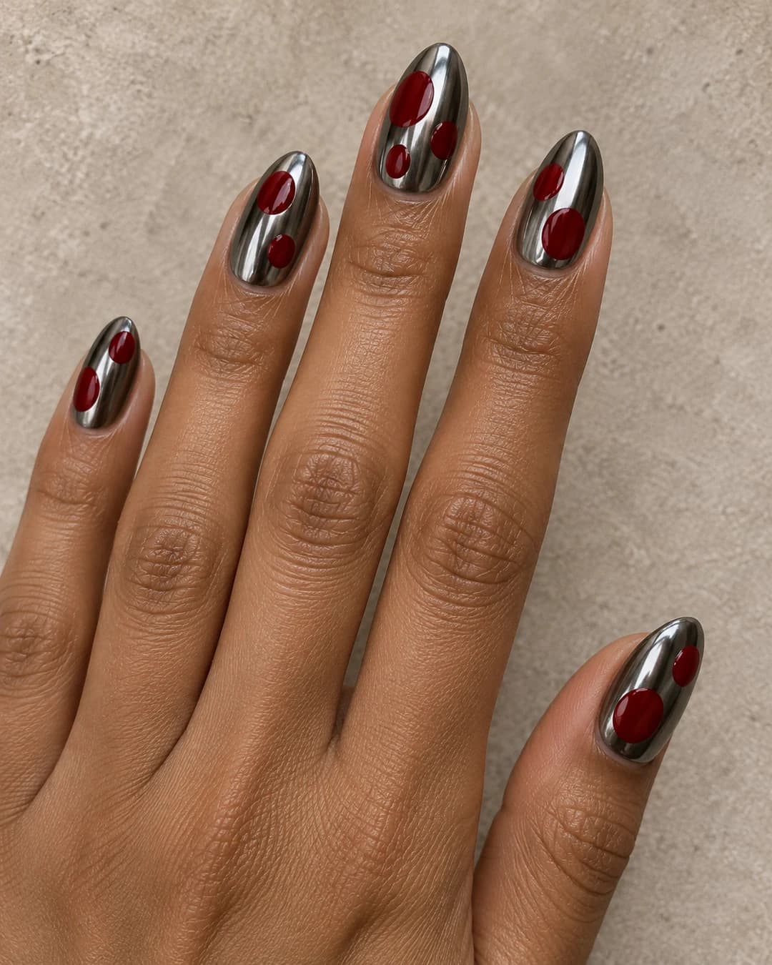 Mano adulta de piel oliva marrón con uñas almendradas medias en cromo gunmetal y lunares cereza grandes de tamaños distintos.