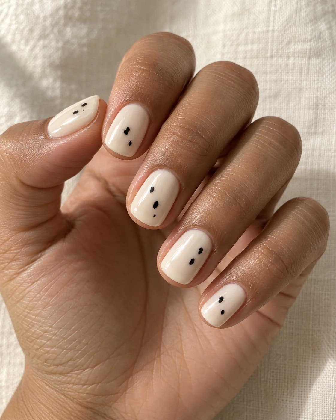 Mano de piel oliva cálida con uñas cortas squoval marfil y pequeños puntos dálmata negros irregulares.