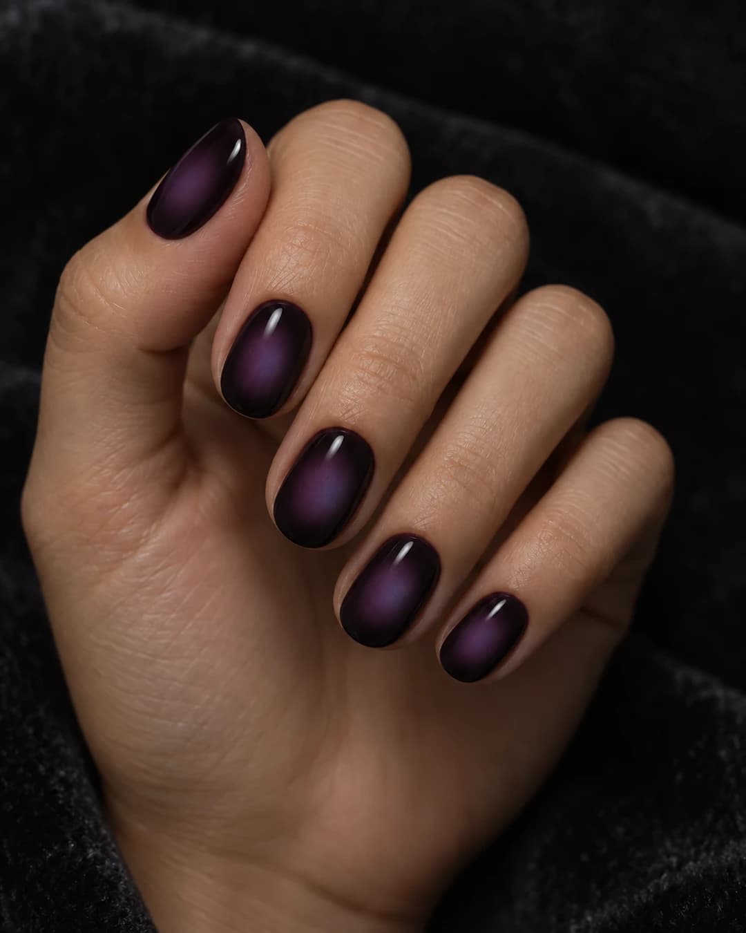 Mano con uñas cortas ovaladas ciruela oscura y un halo difuminado índigo-violeta en cada uña.