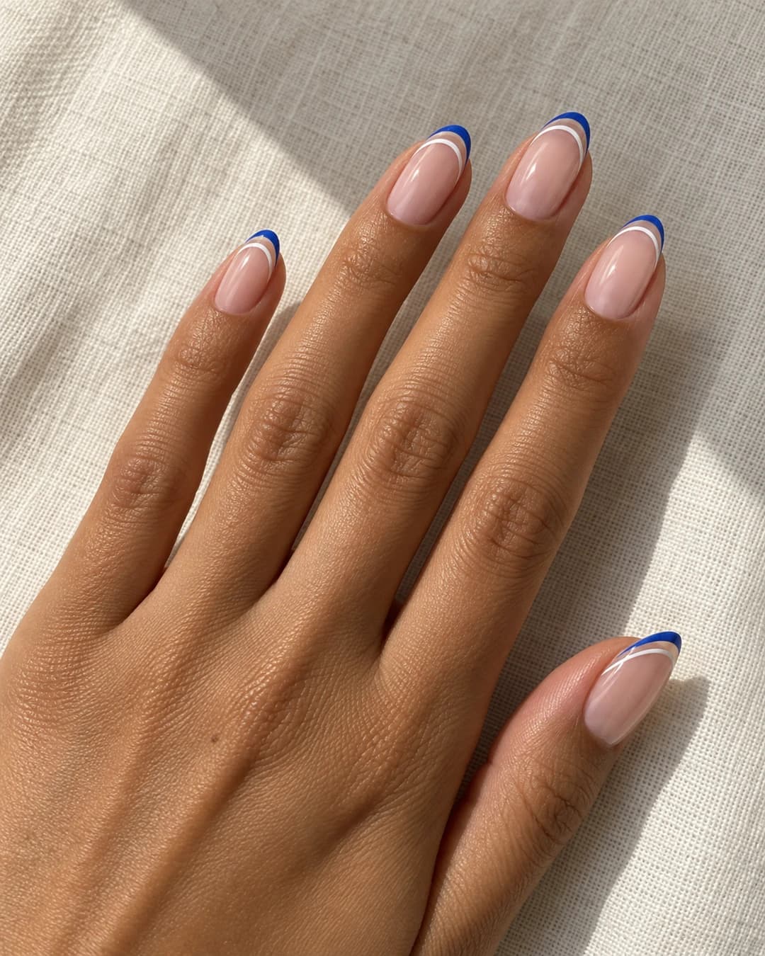 Uñas almendradas medias con dos líneas finas paralelas cobalto y blancas sobre base nude rosa.