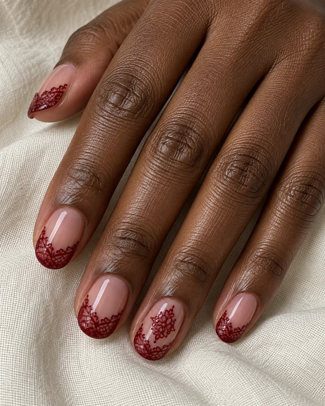 Mano adulta de piel marrón profunda con uñas ovaladas cortas, base rosa translúcida y puntas de encaje burdeos.