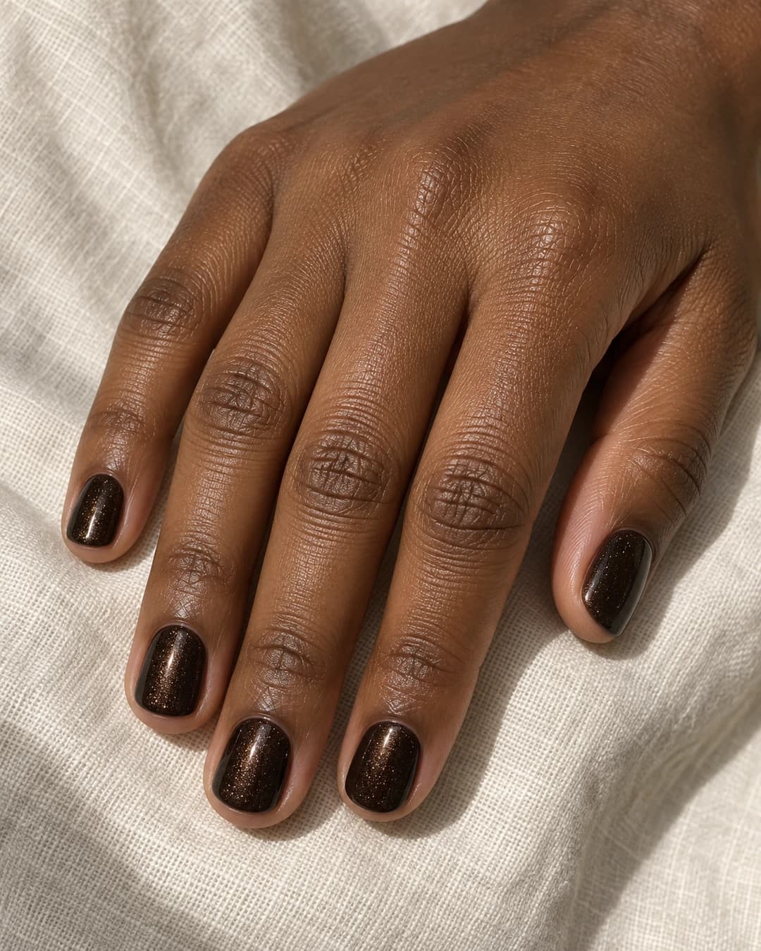 Mano de piel marrón profunda con uñas squoval cortas en chocolate espresso casi negro y microbrillo cálido.