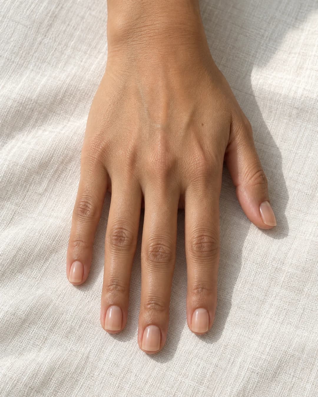 Mano de piel oliva con cinco uñas squoval cortas en nude beige rosado translúcido, sobre lino claro.