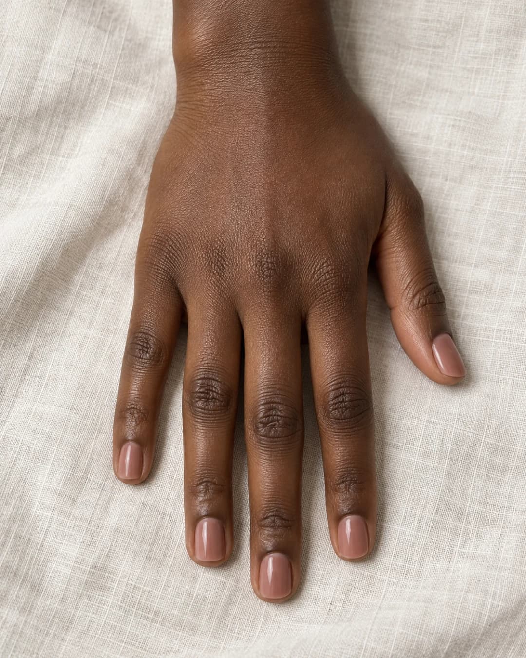 Mano de piel marrón profunda con cinco uñas squoval cortas en nude rosa cacao translúcido, sobre lino claro.