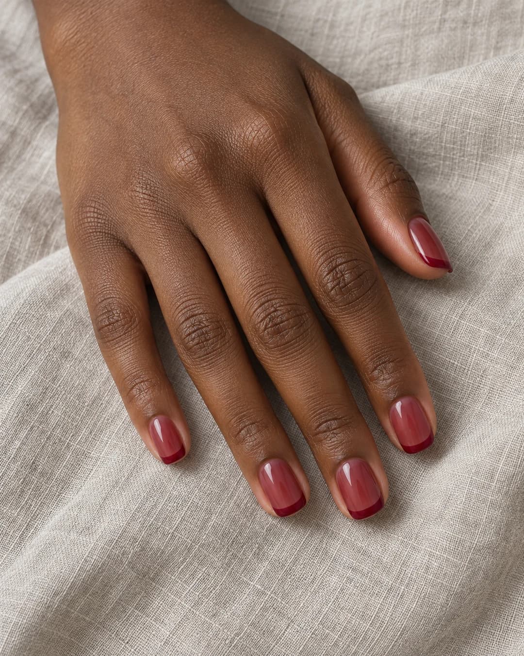 Mano adulta de piel morena profunda con cinco uñas squoval cortas: base jelly frambuesa translúcida y puntas opacas de tono más profundo.