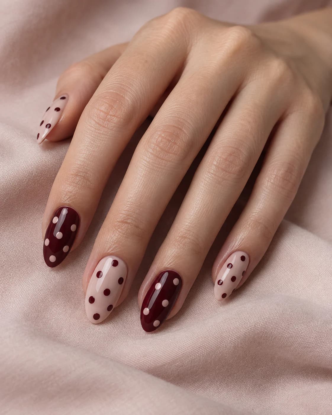Mano adulta de piel clara con cinco uñas almendradas medias que alternan base rosa con puntos vino y base granate con puntos rosa.