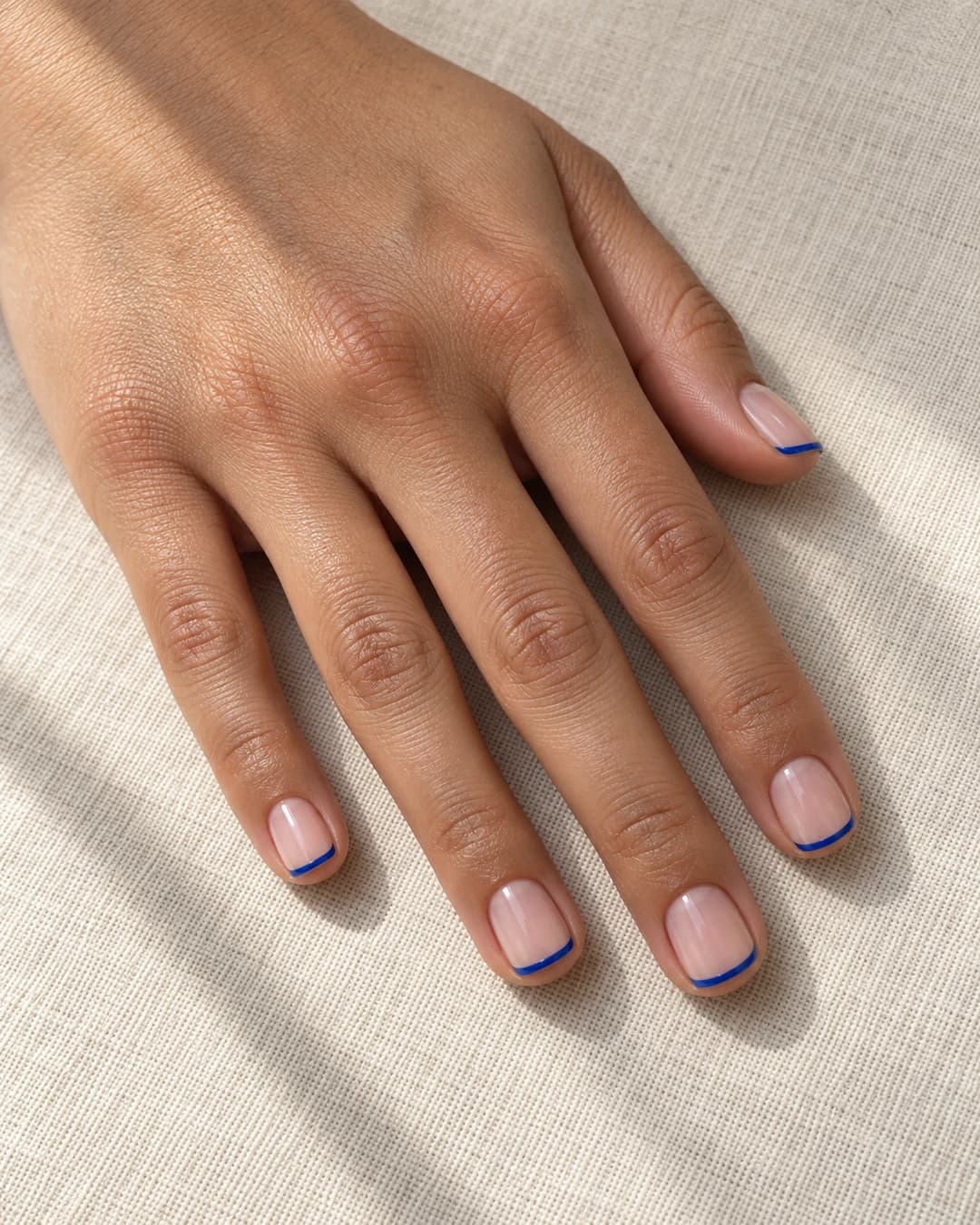 Uñas cortas cuadradas de esquinas suaves con línea micro-french cobalto sobre base nude rosa.
