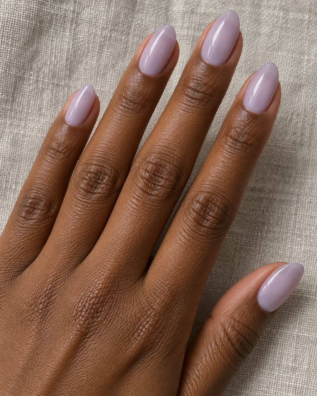 Mano adulta de piel marrón profunda con uñas almendradas medias en lavanda lechoso brillante.