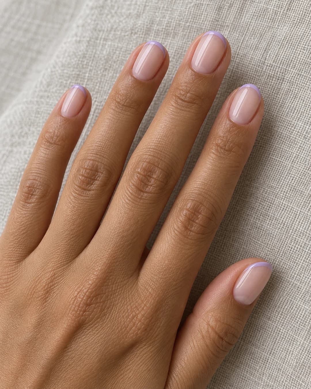 Mano adulta de piel oliva con uñas ovaladas cortas, base rosa lechosa y puntas micro-french lavanda.