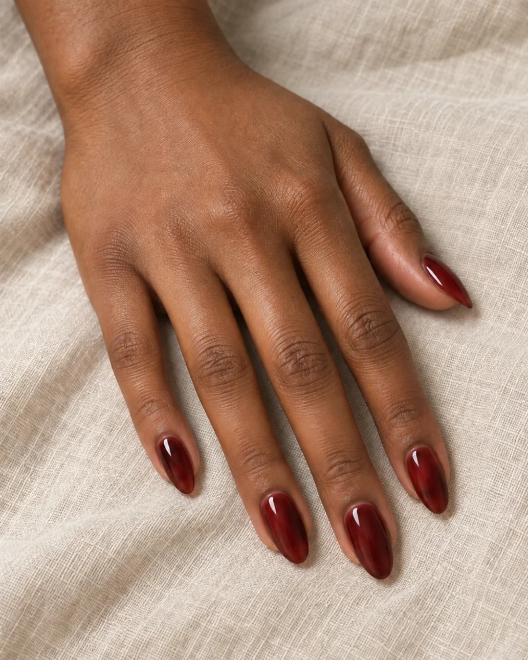 Mano de piel morena con cinco uñas almendradas medias en cherry cola translúcido sobre lino avena.